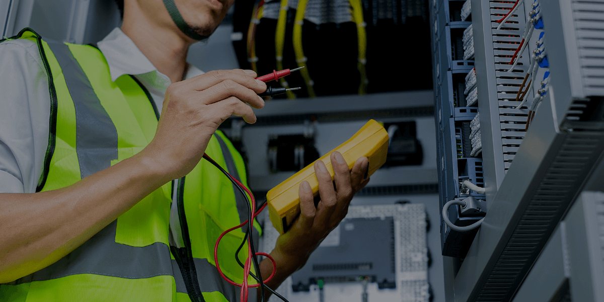 Industrial electrician in high-visibility vest testing electrical panel connections with a digital multimeter.