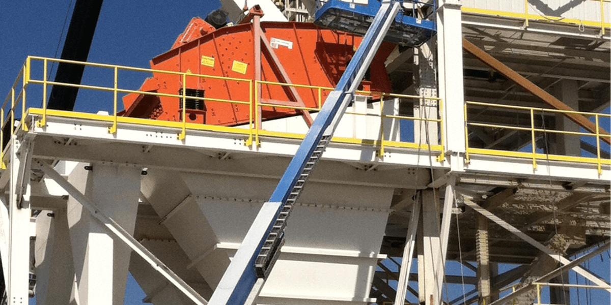 Construction crew working on an elevated industrial structure with conveyors and a bright orange screen tower, using a boom lift for access.