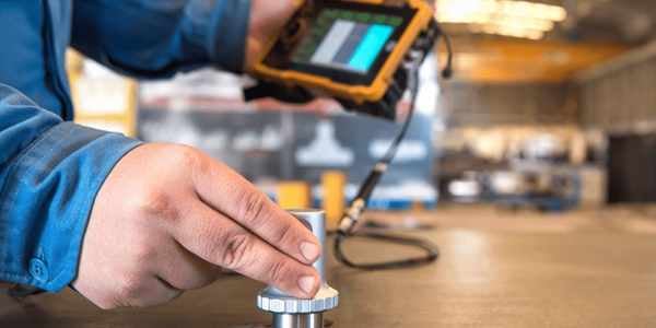 Burkes Mechanical instrumentation technician in a blue work shirt using a handheld ultrasonic calibration device on a metal component inside an industrial facility