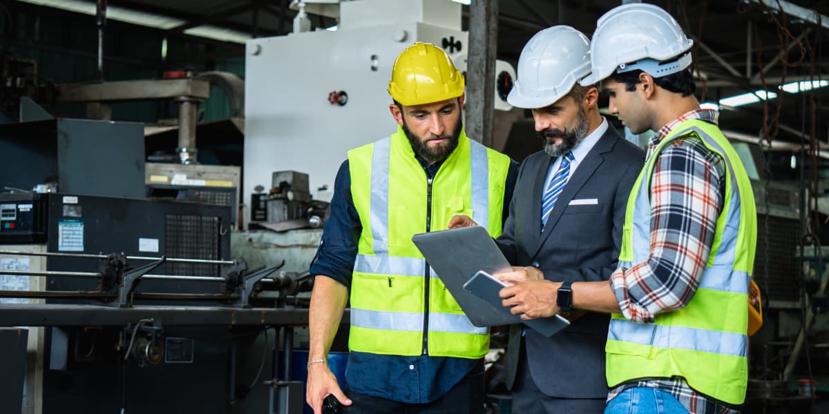 Three Burkes Mechanical industrial team members wearing hard hats and high visibility vests reviewing a project work order on a tablet inside an active manufacturing facility
