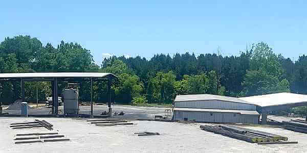 Burkes Mechanical outdoor fabrication and industrial coatings yard with structural steel materials staged on the ground, open-air covered work area, and metal shop buildings surrounded by trees