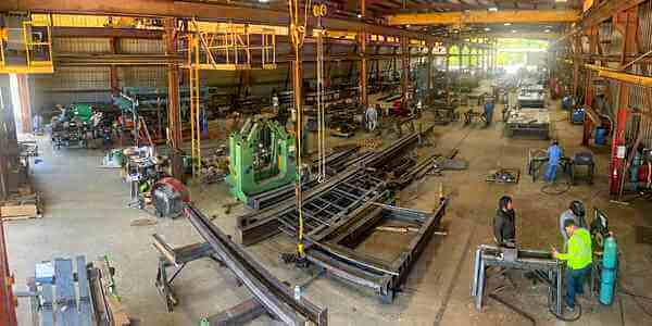 Burkes Mechanical structural steel fabrication shop with overhead cranes, workers in safety vests assembling large steel frames, and multiple active workstations across a spacious industrial facility